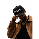Person wearing a black cap with 'FOUND' text, brown jacket, and gold chains on a white background