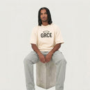 Person wearing a cream t-shirt with 'GRCE' logo sitting on a concrete block against a plain background